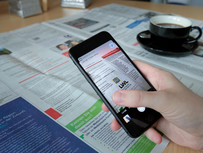 Ein Handy zeigt auf den CR-Code einer Ausschreibung in der Zeitung.