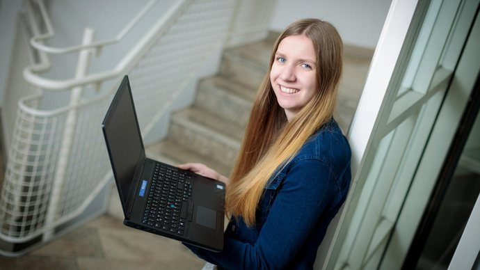 Eine Studentin des Fachbereichs Bachelor of Science Informatik lehnt mit einem Notebook in der Hand an einer Säule und lacht in die Kamera